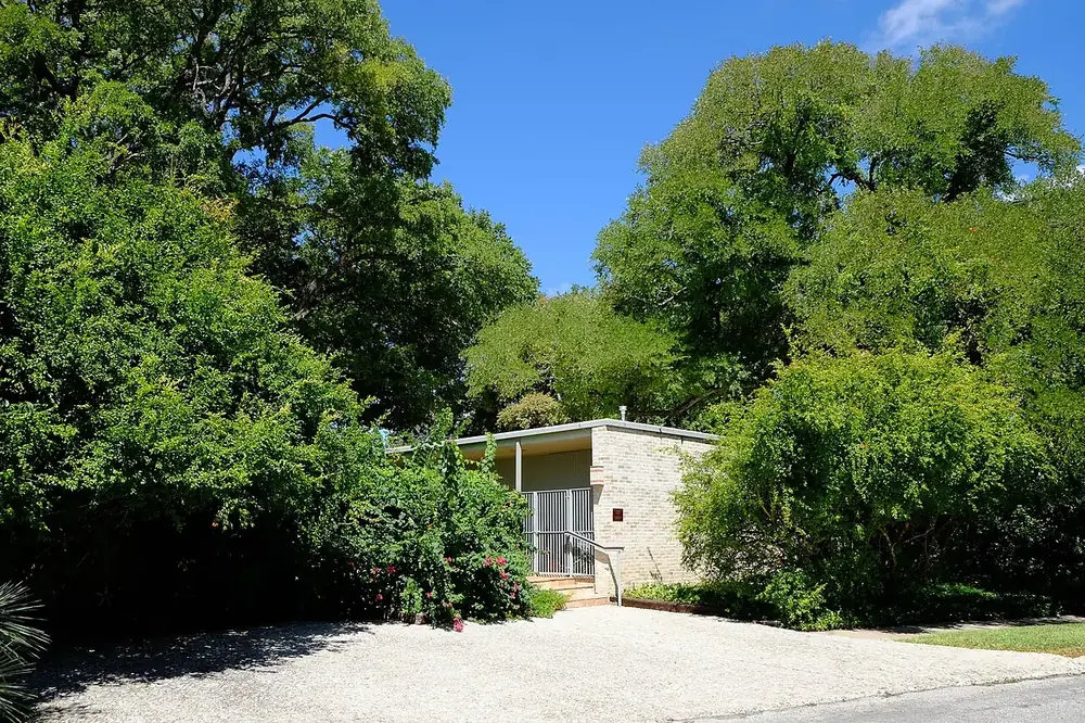 A low mid-century house in Alamo Heights set behind big shade trees and shrubs